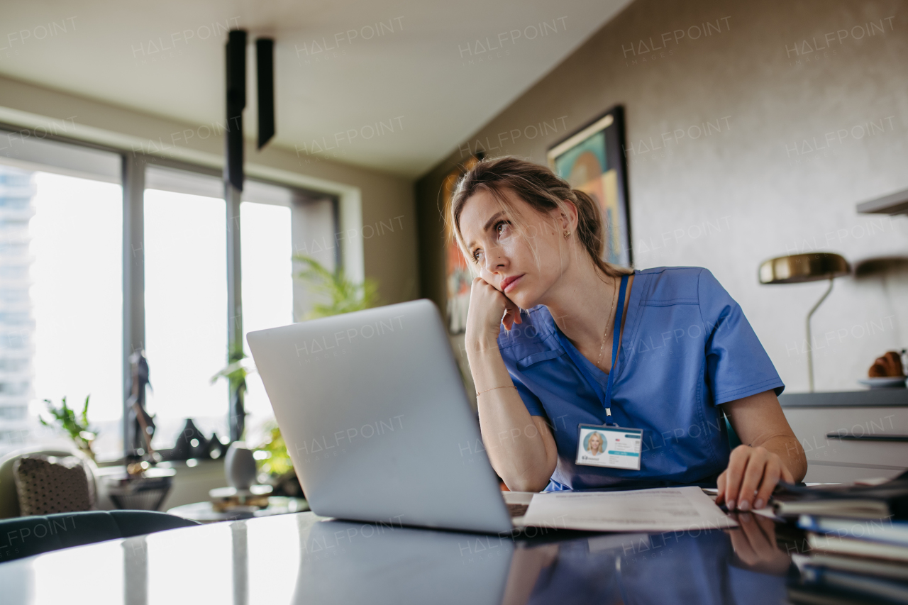 Nurse paying bills online, sitting in living room, working on notebook. Doctor in scrubs dealing with work issues from home, after workday, reading medical record, telemedicine consultation. Work-life balance for healthcare worker.