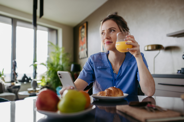 Female nurse or doctor getting ready for work in the morning, scrolling on smartphone while eating breakfast, drinking juice. Work-life balance for healthcare worker.