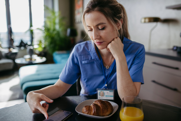 Female nurse or doctor getting ready for work in the morning, scrolling on smartphone while eating breakfast, before leaving for the work dressed in scrubs. Work-life balance for healthcare worker.