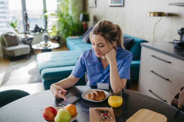 Female nurse or doctor getting ready for work in the morning, scrolling on smartphone while eating breakfast, before leaving for the work dressed in scrubs. Work-life balance for healthcare worker.