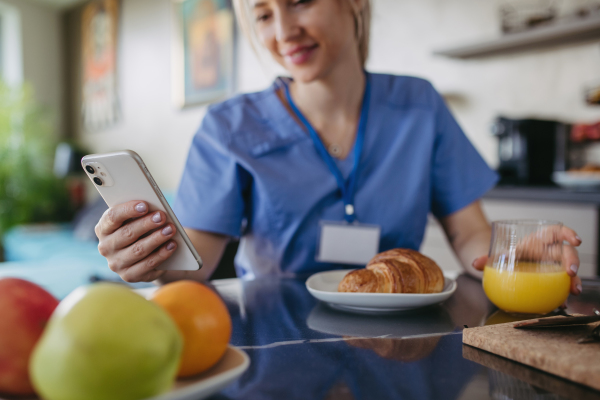 Female nurse or doctor getting ready for work in the morning, scrolling on smartphone while eating breakfast, before leaving for the work dressed in scrubs. Work-life balance for healthcare worker.