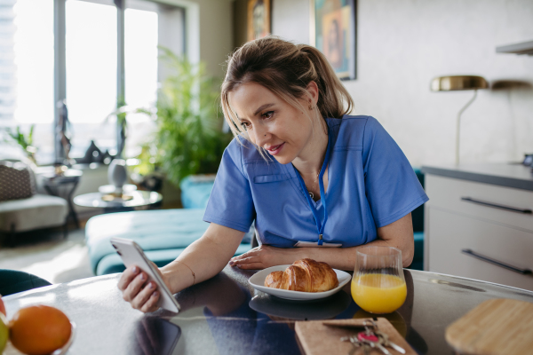 Female nurse or doctor getting ready for work in the morning, scrolling on smartphone while eating breakfast, before leaving for the work dressed in scrubs. Work-life balance for healthcare worker.