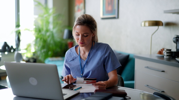 Nurse paying bills online, sitting in living room, working on notebook. Doctor in scrubs dealing with work issues from home, after workday, reading medical record, telemedicine consultation. Work-life balance for healthcare worker.