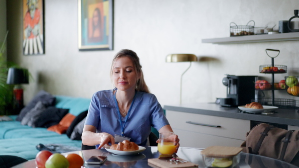 Female nurse or doctor getting ready for work in the morning, scrolling on smartphone while eating breakfast, before leaving for the work dressed in scrubs. Work-life balance for healthcare worker.