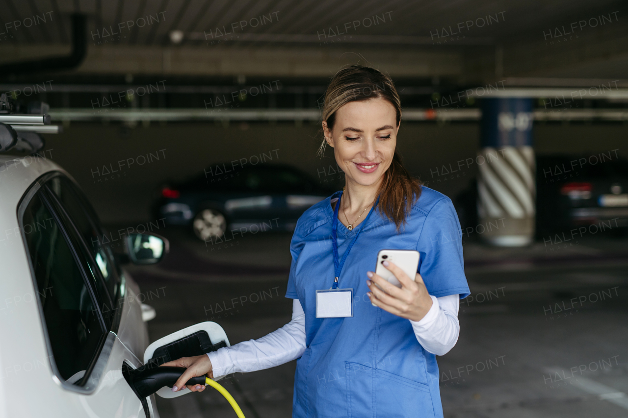 Nurse plugging charger into her electric car, before going to work. Electric vehicle charging station in front of hospital, modern clinic. Charging at work, workplace.