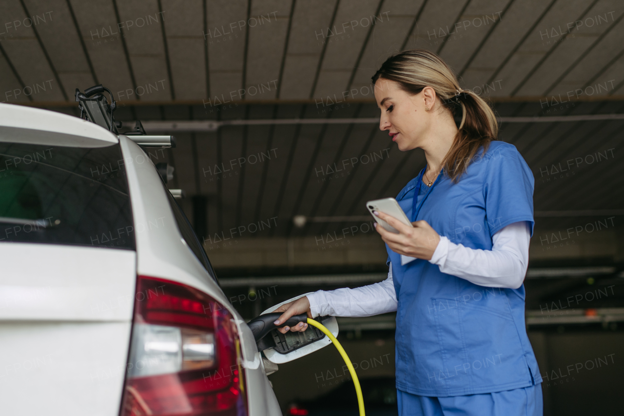 Nurse plugging charger into her electric car, before going to work. Electric vehicle charging station in front of hospital, modern clinic. Charging at work, workplace.