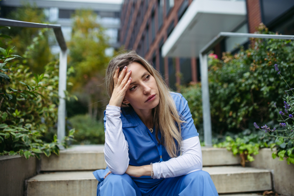 Young female doctor feeling overwhelmed at work, sitting on concrete stairs, looking at phone. Healthcare workers having stressful job, feeling exhausted. Burnout syndrome for doctors and nurses.