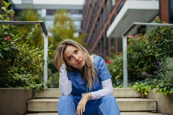 Young female doctor feeling overwhelmed at work, sitting on concrete stairs, looking at phone. Healthcare workers having stressful job, feeling exhausted. Burnout syndrome for doctors and nurses.