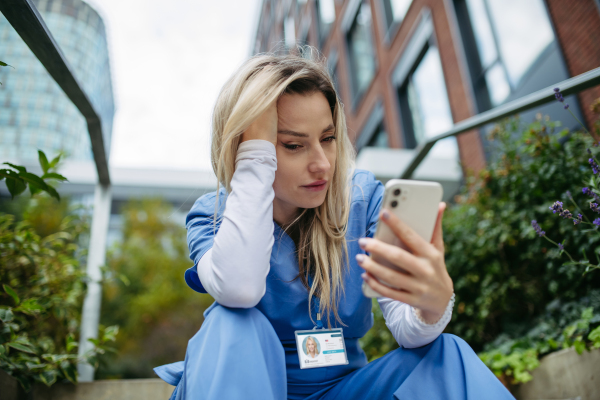 Young female doctor feeling overwhelmed at work, sitting on concrete stairs, looking at phone. Healthcare workers having stressful job, feeling exhausted. Burnout syndrome for doctors and nurses.