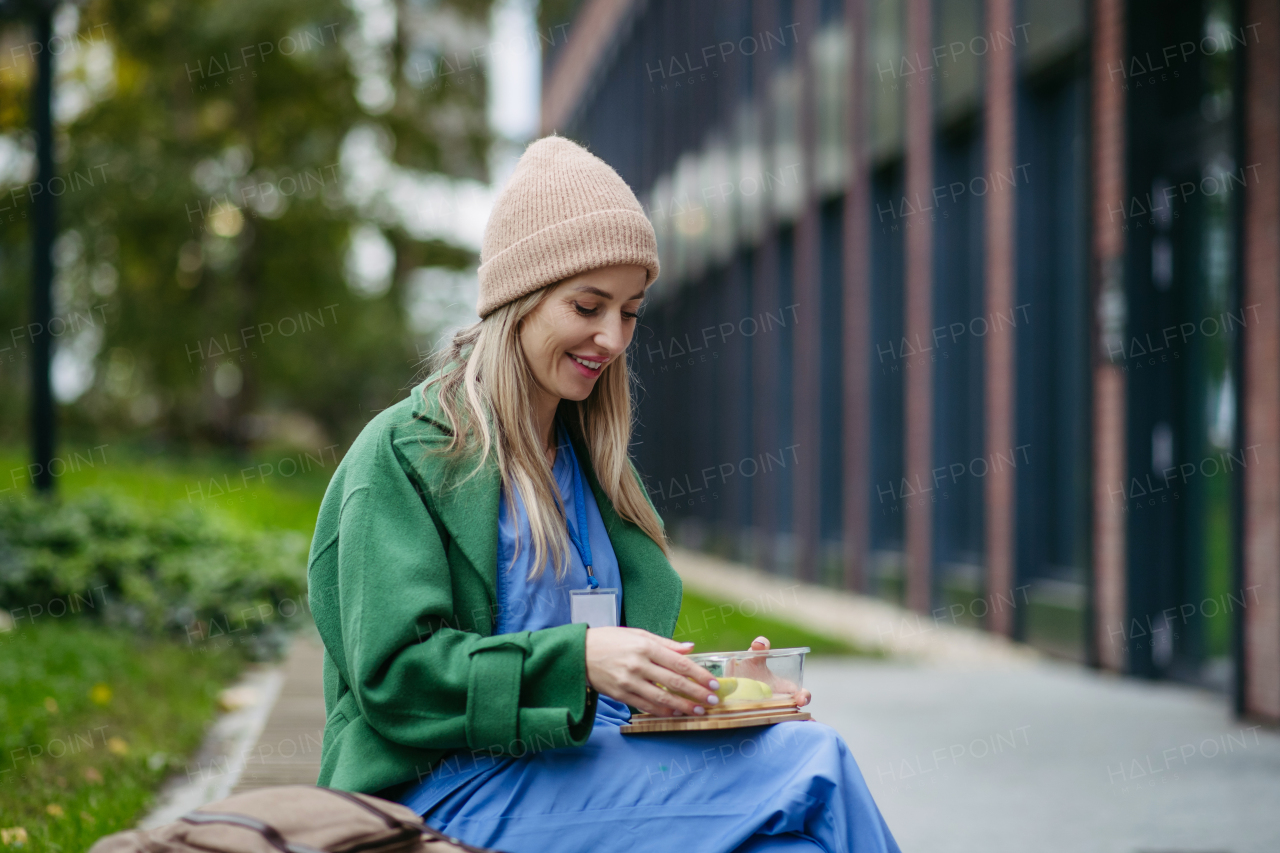 Nurse having healthy balanced diet, fruit snack in front of hospital building, taking break from work. Importance of breaks in healthcare.