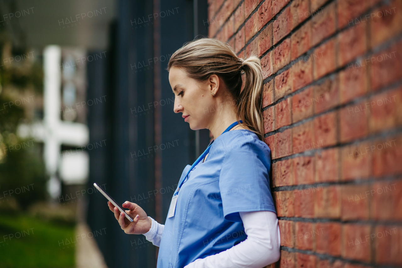 Nurse, doctor in blue uniform taking break, leaning on hospital building, scrolling on smartphone. Work-life balance of healthcare worker.