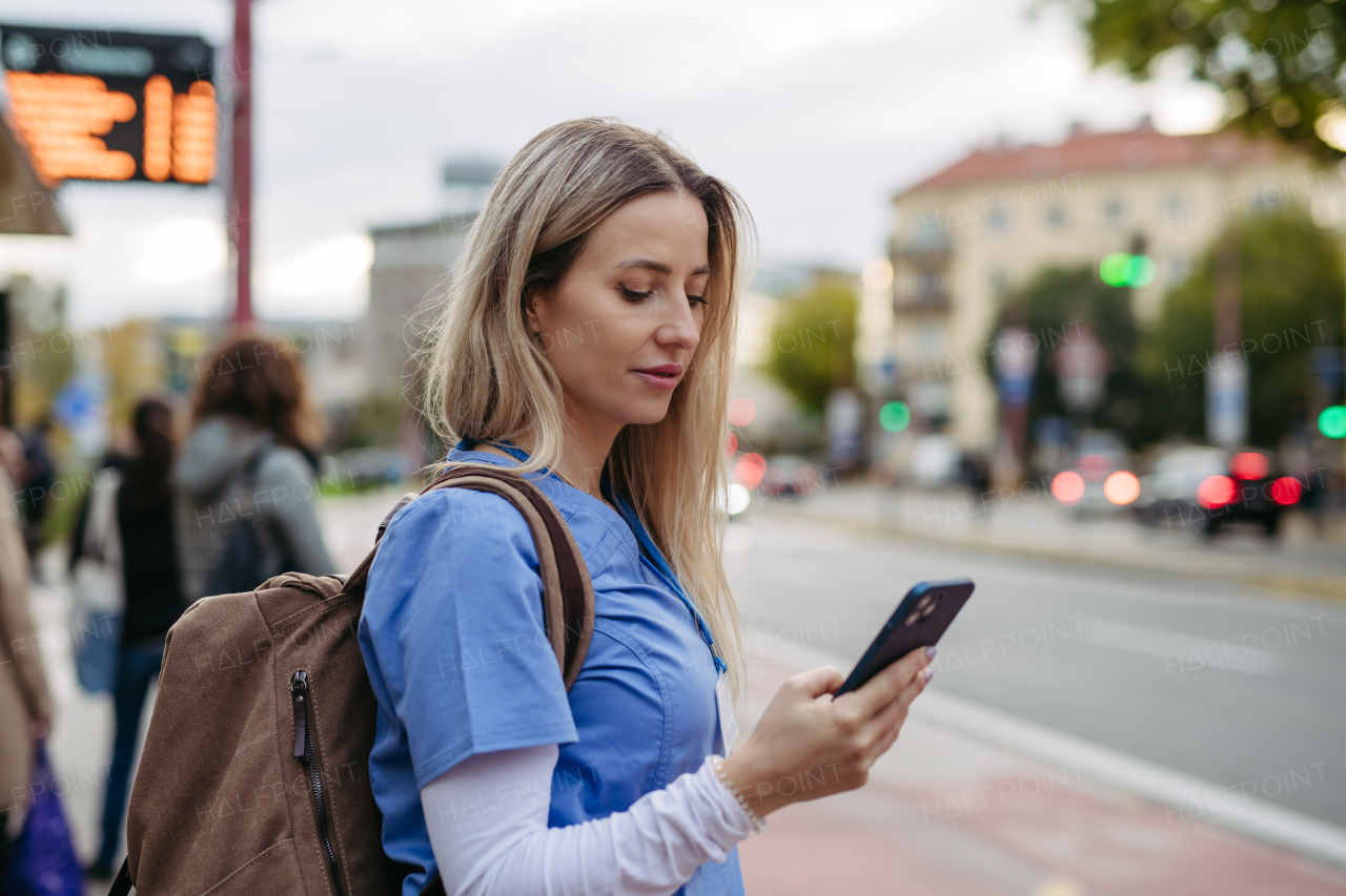 Female nurse, doctor going home from work, waiting for bus, scrolling on smartphone. Work-life balance of healthcare worker.