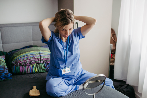 Female nurse or doctor getting ready for work in the morning, brushing hair, making ponytail in bedroom, dressed in scrubs. Work-life balance for healthcare worker.