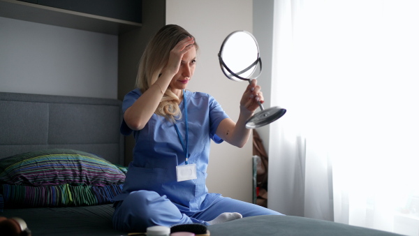 Female nurse or doctor getting ready for work in the morning, doing makeup, skincare in bedroom, dressed in scrubs. Work-life balance for healthcare worker.