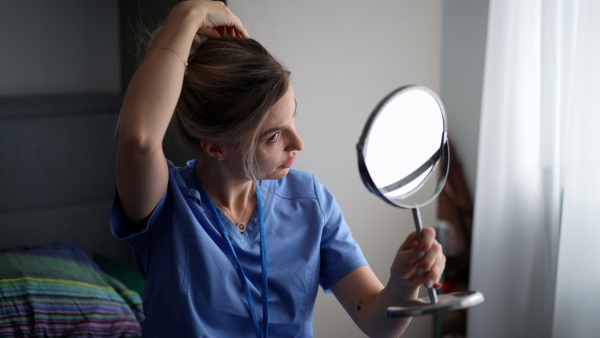 Female nurse or doctor getting ready for work in the morning, doing makeup, skincare in bedroom, dressed in scrubs. Work-life balance for healthcare worker.