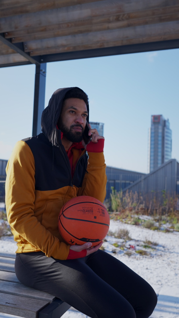 Young multiracial man sitting outdoor in a city with basketball. Vertical view.