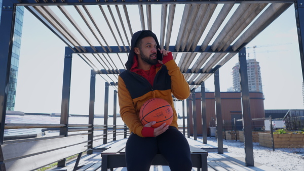 Young multiracial man sitting outdoor in a city with basketball.