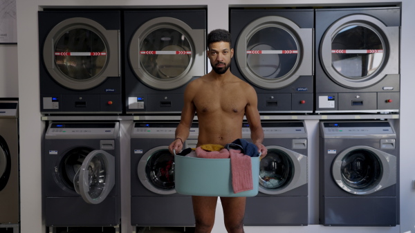 Young naked multiracial man in laundry, holding laundry basket.