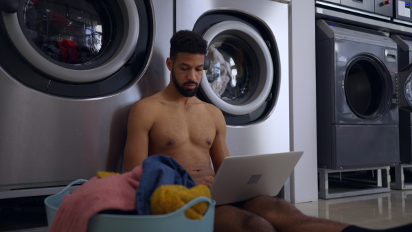 Young naked multiracial man in laundry, sitting on the floor, waiting with laptop.