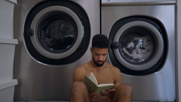 Young naked multiracial man in laundry, sitting on the floor, waiting and reading book.