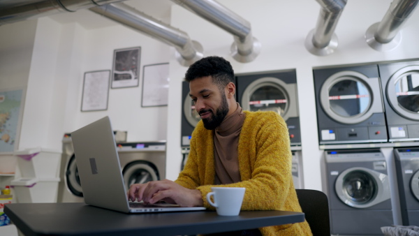 Young multiracial man in laundry working on laptop, drinking coffee and waiting.