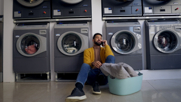 Young multiracial man in laundry, sitting on the floor, calling and waiting.