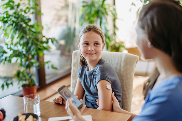 Girl with diabetes checking blood glucose level at home using continuous glucose monitor. Girl's mother connects CGM to a smartphone to monitor her blood sugar levels in real time.