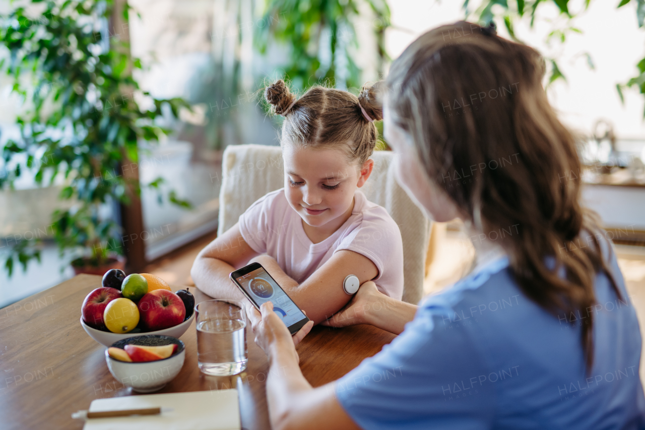 Girl with diabetes checking blood glucose level at home using continuous glucose monitor. Girl's mother connects CGM to a smartphone to monitor her blood sugar levels in real time.