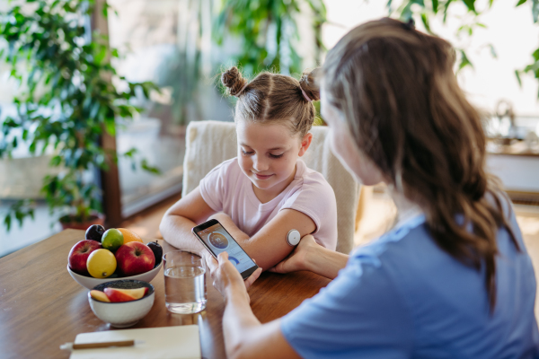 Girl with diabetes checking blood glucose level at home using continuous glucose monitor. Girl's mother connects CGM to a smartphone to monitor her blood sugar levels in real time.