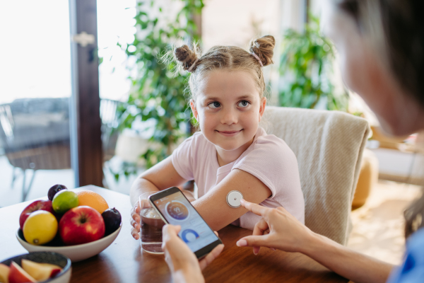 Girl with diabetes checking blood glucose level at home using continuous glucose monitor. Girl's mother connects CGM to a smartphone to monitor her blood sugar levels in real time.