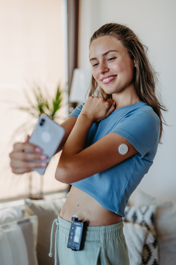 Beautiful diabetic girl taking a selfie, having an insulin pump and a continuous glucose monitor on her arm. Young woman is not ashamed of having diabetes, sharing her life with a chronic illness on social media.