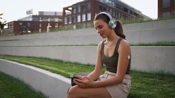 Diabetic woman with continuous glucose monitor sitting outdoors in park, watching social media content on smart phone.The CGM device makes the life of the schoolgirl. easier, helping manage his illness and focus on other activities.