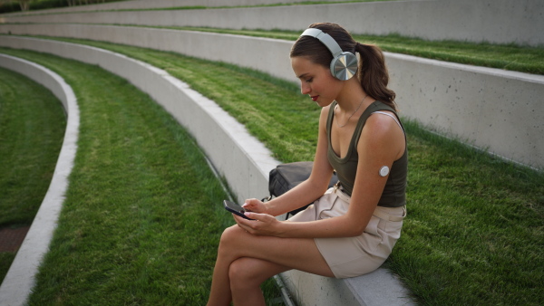 Diabetic woman with continuous glucose monitor sitting outdoors in park, watching social media content on smart phone.The CGM device makes the life of the schoolgirl. easier, helping manage his illness and focus on other activities.