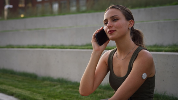 Diabetic woman with continuous glucose monitor sitting outdoors in park, having a call outside in the city park. The CGM device makes the life of the schoolgirl. easier, helping manage his illness and focus on other activities.