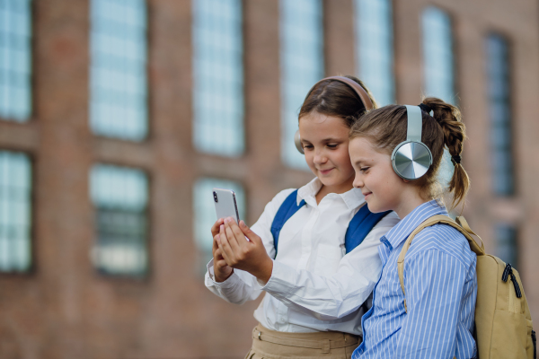 Two schoolgirls are checking social media on their smartphone in front of the school. Friends with headphones are watching a video.