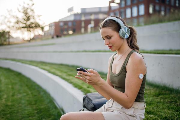 Diabetic woman with continuous glucose monitor sitting outdoors in park, watching social media content on smart phone.The CGM device makes the life of the schoolgirl. easier, helping manage his illness and focus on other activities.