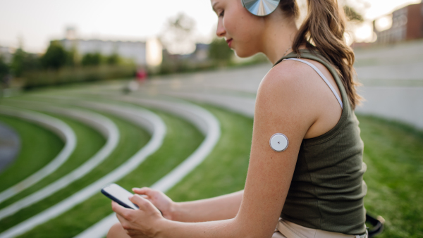 Diabetic woman with continuous glucose monitor sitting outdoors in park, watching social media content on smart phone.The CGM device makes the life of the schoolgirl. easier, helping manage his illness and focus on other activities.