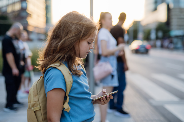 Side view of a girl waiting at the pedestrian crossing, scrolling on her smartphone on her way to school. Concept of back to school.