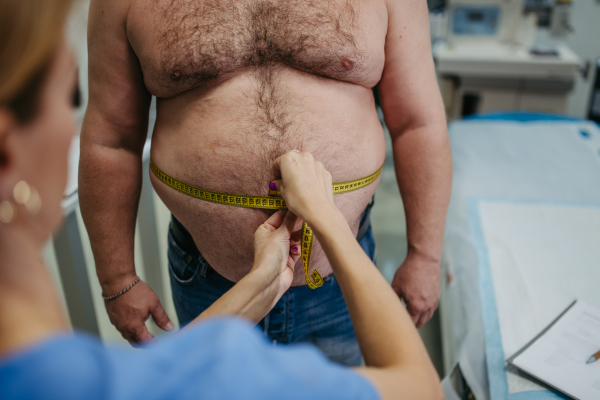 Female doctor measuring waist of overweight patient using a tape measure. Obesity affecting middle-aged men's health. Concept of health risks of overwight and obesity.