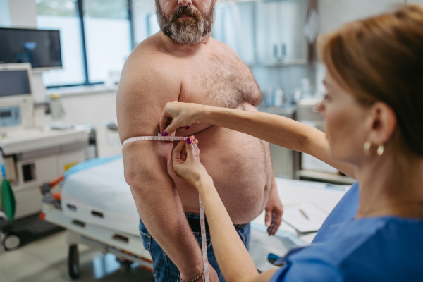 Female doctor measuring arm circumference of overweight patient using a tape measure. Obesity affecting middle-aged men's health. Concept of health risks of overwight and obesity.