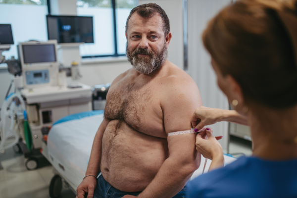 Female doctor measuring arm circumference of overweight patient using a tape measure. Obesity affecting middle-aged men's health. Concept of health risks of overwight and obesity.