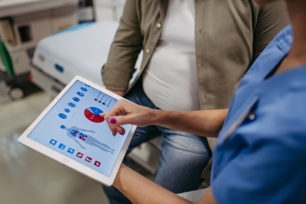 Close up of doctor consulting with the overweight patient, discussing test result in doctor office. Showing medical report on tablet. Obesity affecting middle-aged men's health. Concept of health risks of overwight and obesity.