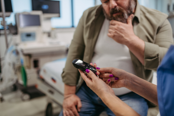 Doctor checking blood glucose level using a fingerstick glucose meter, waiting for results from glucometer. Obese or overweight man at risk of developing type 2 diabetes. Concept of health risks of overwight and obesity.