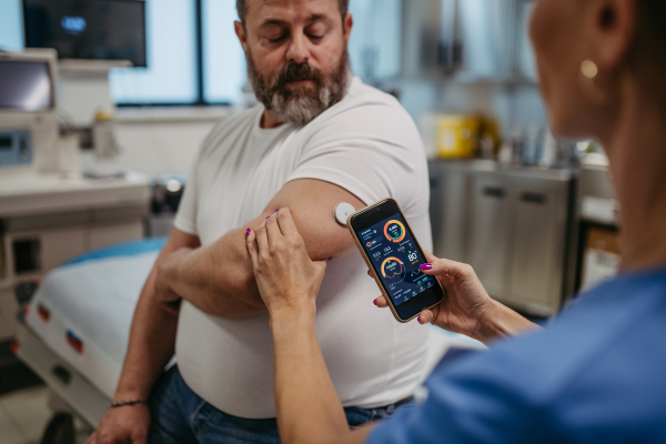 Doctor connecting patient's continuous glucose monitor with smartphone, to check his blood sugar level in real time. Obese, overweight man is at risk of developing type 2 diabetes. Concept of health risks of overwight and obesity.