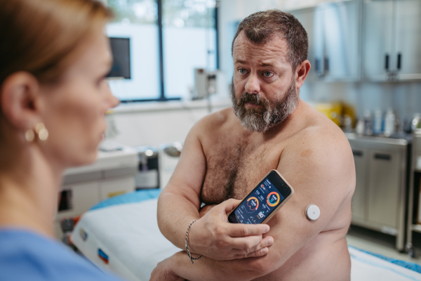 Doctor connecting patient's continuous glucose monitor with smartphone, to check his blood sugar level in real time. Obese, overweight man is at risk of developing type 2 diabetes. Concept of health risks of overwight and obesity.