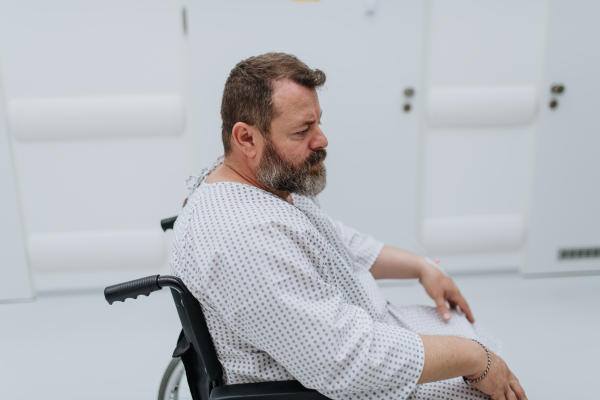 Side view of the man in wheelchair. Overweight patient in hospital gown waiting for medical examination in hospital, feeling anxious.