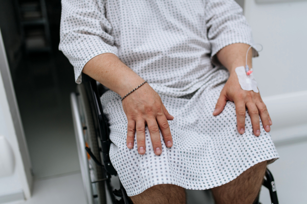 Close up of man in wheelchair with IV cannula in hand. Overweight patient in hospital gown waiting for medical examination in the hospital.