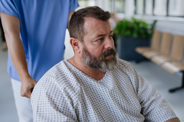 Male nurse pushing a patient in wheelchair along a hospital corridor. Overweight patient feeling anxious and has health concerns.