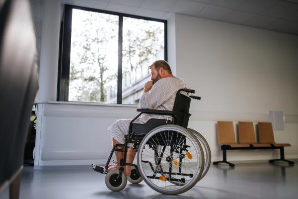 Side view of the man in wheelchair. Overweight patient in hospital gown waiting for medical examination in hospital.