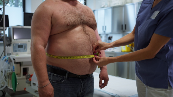 Female doctor measuring waist of overweight patient using a tape measure. Obesity affecting middle-aged men's health. Concept of health risks of overwight and obesity.